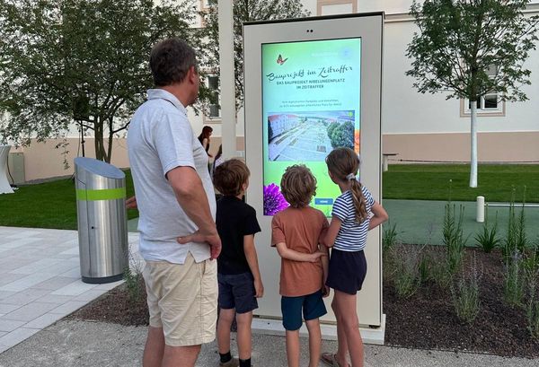 Kinder und Mann stehen vor digitalen Stele und schauen dem Bauvideo zu